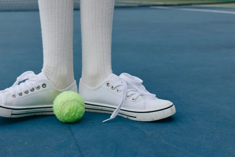 Young athlete in white sneakers and long socks playing tennis on a blue court with a green ball, showcasing passion and determination in sports