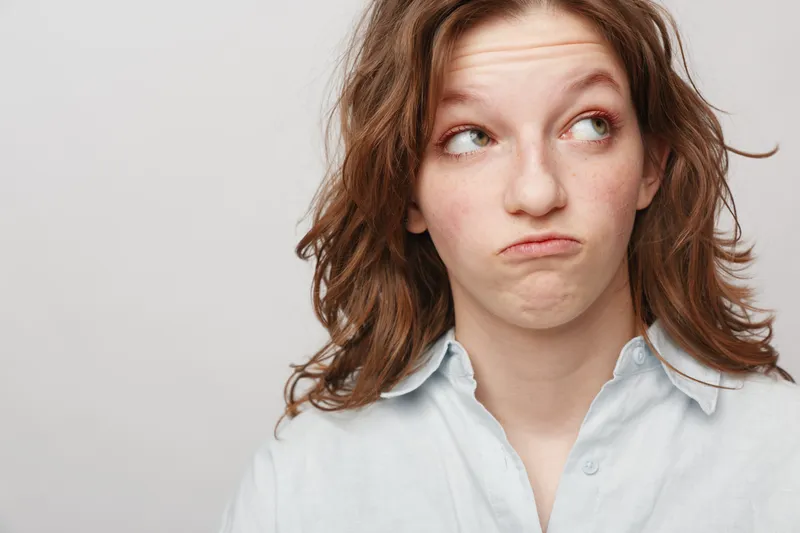 Young woman with red hair and freckles looks confused and skeptical wearing a white shirt on a plain light background. Concept of doubt, uncertainty, and contemplation.