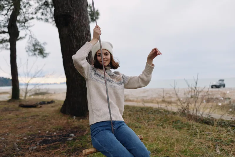 Woman on a rope swing by a tree at a sandy beach, wearing a cozy knit sweater and beanie, enjoying a sunny day, capturing carefree outdoor recreation and nature vibes.