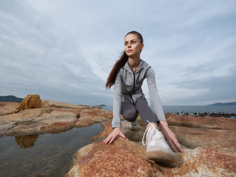 Young athletic woman stretching on rocky beach during cloudy day, wearing grey sportswear, embodying fitness and wellness