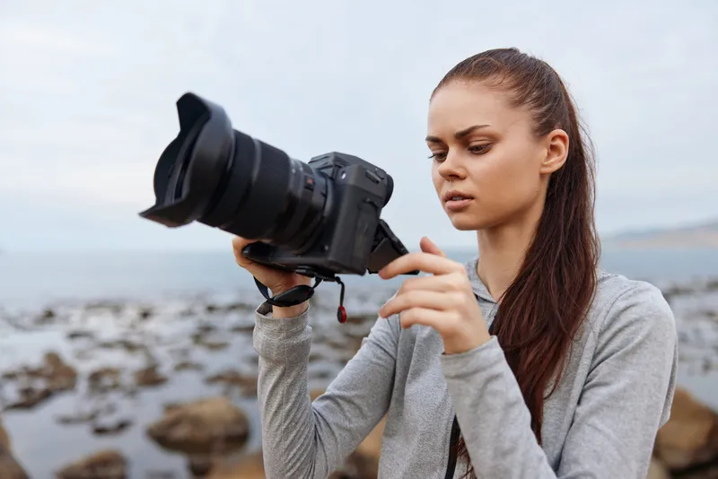 Young woman using a professional camera on a rocky beach, wearing casual grey sportswear, capturing the beauty of nature with a focused expression