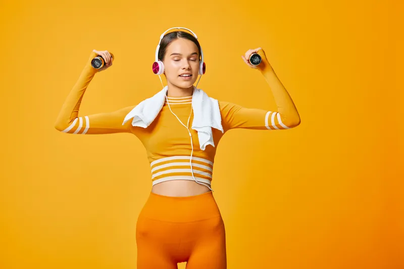 Fitness, woman workout in a vibrant orange outfit, lifting dumbbells with a towel and headphones, high-energy studio vibe and bold motion against a bright background.