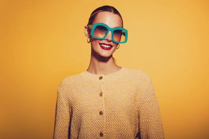 Smiling woman in yellow knitted sweater wears large turquoise sunglasses, posing against bright yellow background. Concept of fashion, style, confidence, and vibrant colors in studio.