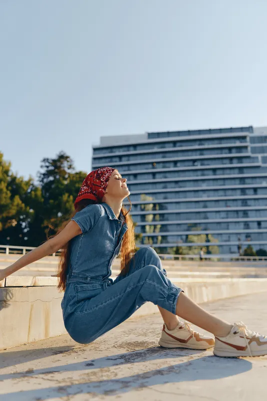 Lifestyle woman wearing modern boho-western denim street style outfit and red bandana, relaxing outdoors in warm natural sunlight, film color effect.
