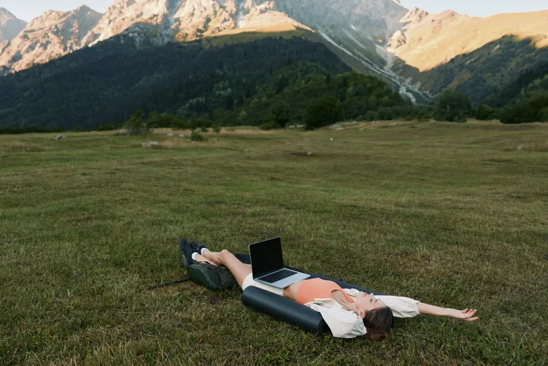A person lies on a grassy meadow with a laptop, enjoying a relaxed outdoor workspace amid rolling hills and distant peaks under bright daylight.