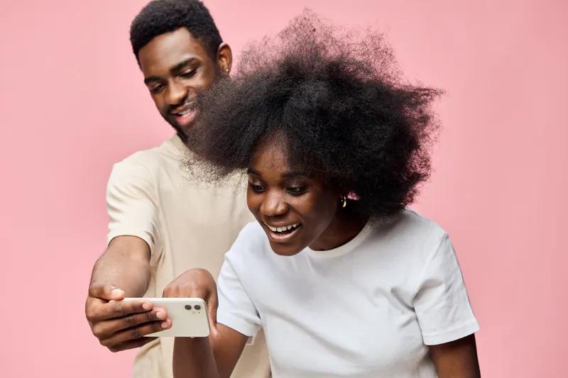 Happy couple enjoying time together, looking at smartphone, with cheerful expressions, against a soft pink background, perfect for social moments and connection