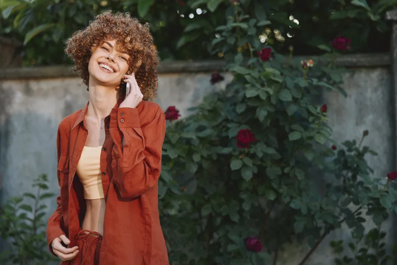 Young woman with curly hair smiles happily, wearing an orange jacket over a yellow crop top in a blooming garden, capturing a casual outdoor portrait with warm, carefree mood and soft sunlight