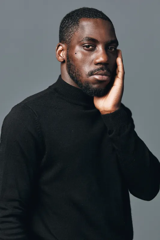 African man with dark skin and beard wearing black turtleneck sweater posing with hand on face in creative conceptual visual style studio portrait on gray background.
