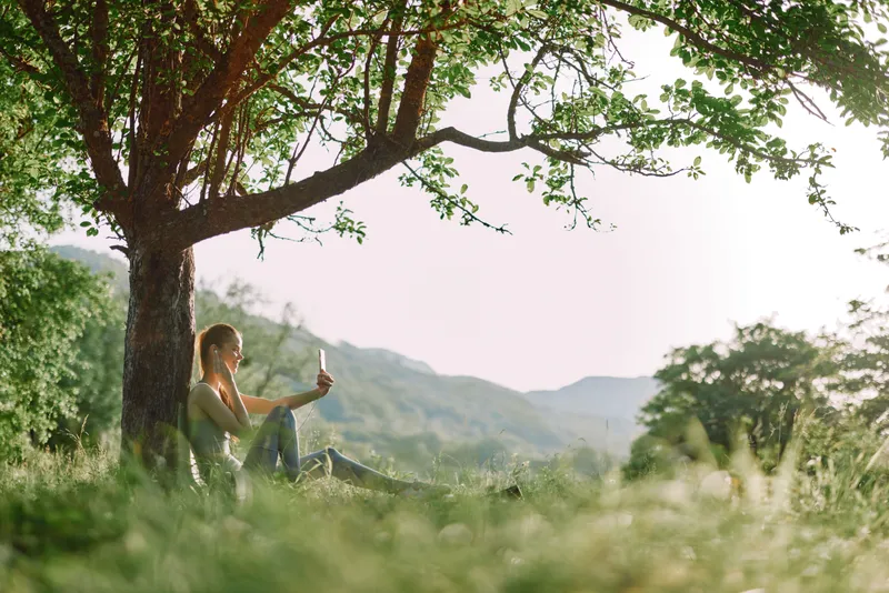 Young woman enjoying a drink under a tree in a sunny natural setting, smiling and relaxed, embodies a healthy lifestyle and harmony with nature