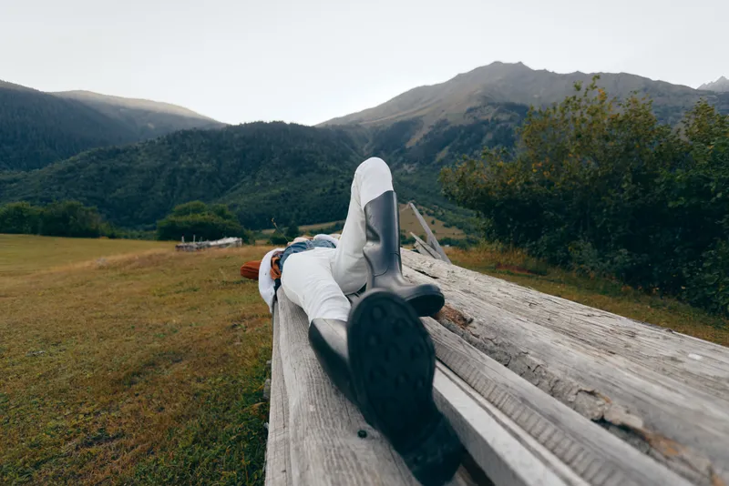 Person lying on a wooden bench in mountains and nature, wearing boots and casual jacket, outdoors relaxation and rest in a peaceful meadow landscape with panoramic horizon.
