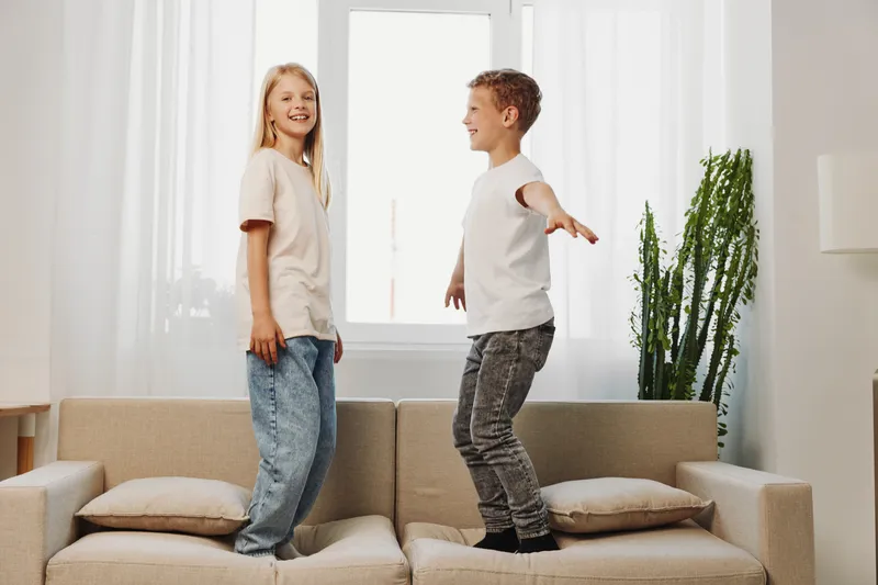 Happy children jumping on sofa in bright living room with large window and green plant, enjoying playful moment and indoor fun at home.