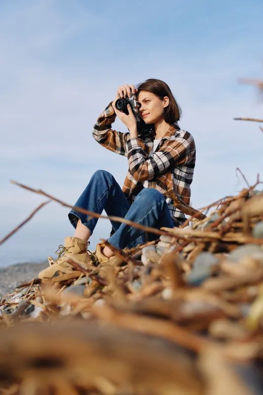 Female photographer perched among autumnal debris, wearing plaid shirt and jeans, aiming a camera toward the landscape with coastal breeze, bright sky, distant horizon, and rugged foreground of dried