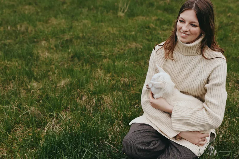 Woman outdoors, sitting on grass, gently holding a goat kid, cozy cream sweater, soft sunlight, peaceful moment, natural colors, simple rural lifestyle, in a quiet meadow with animal companionship