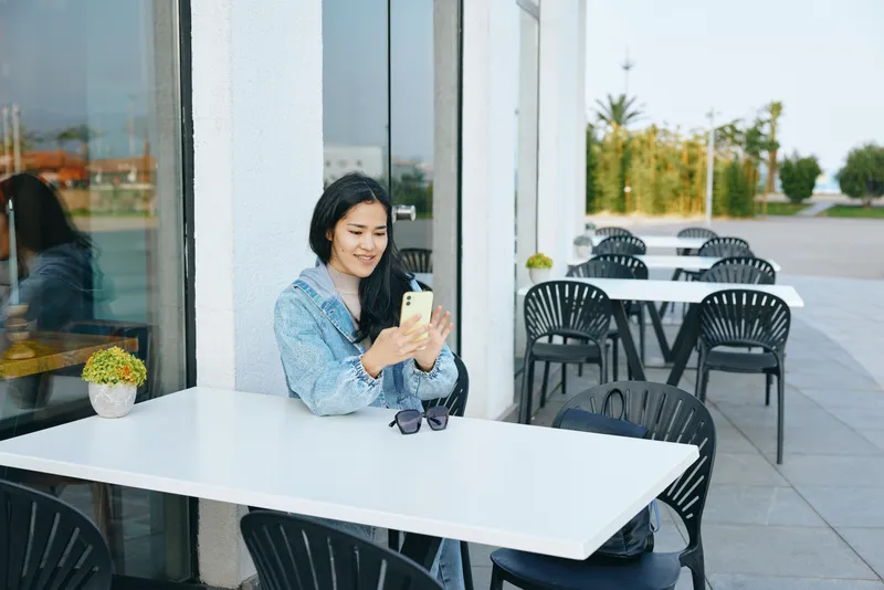Female at outdoor cafe uses smartphone, sitting at a white table, sunglasses beside, black chairs, bright day, relaxed urban atmosphere, casual dining