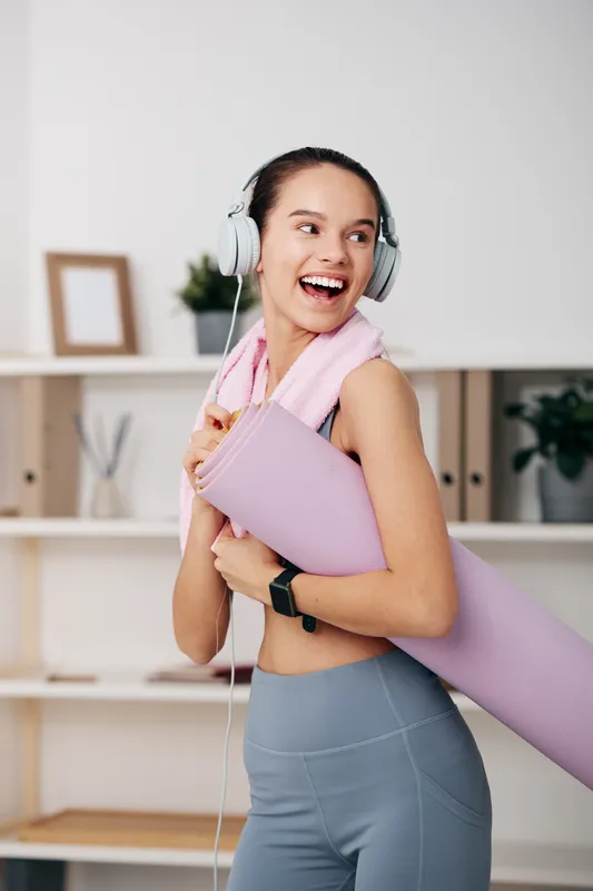 Fitness woman wearing headphones, carrying a yoga mat over her shoulder, smiling in a bright home gym with shelves and plants.