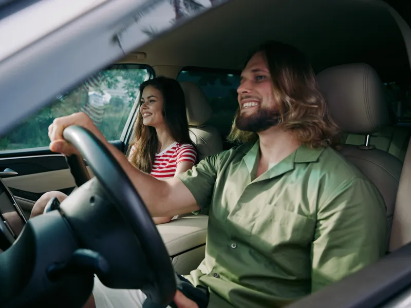 Two friends enjoy a sunny car ride; the man drives with a wide smile while the woman chats beside him, creating a bright, relaxed travel moment inside the cabin.