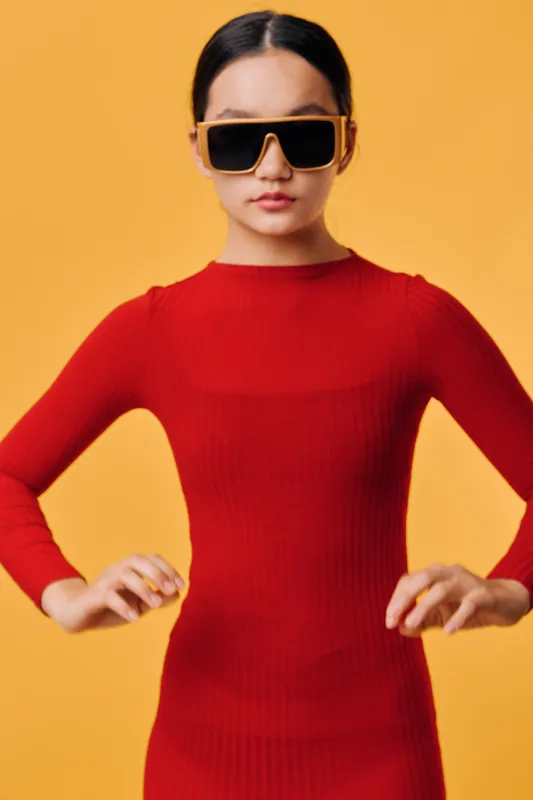 Woman in sunglasses and red dress standing with hands on hips against yellow background, fashion portrait with confident pose and modern style studio shot.