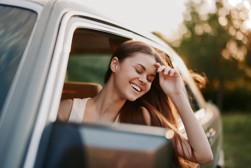 Young woman smiling joyfully from a car window, enjoying a sunny day, showcasing carefree emotions and a sense of freedom in a natural green background.
