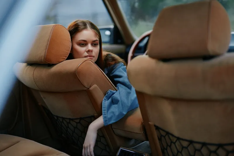 A young woman with long brown hair in a denim jacket looks thoughtfully through the back seat of a vintage car, evoking a sense of contemplation and nostalgia.