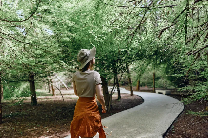 Woman walking on winding path through forest with hat, trees and green nature, casual stroll on shaded trail, back view of traveler with sweater tied around waist enjoying peaceful outdoor escape.