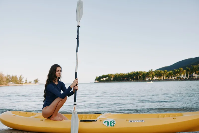 A woman sits on a bright yellow kayak, gripping a double blade paddle, in calm water under a clear sky, poised for a steady paddle stroke.