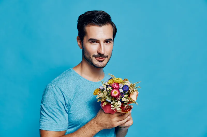 Handsome smiling man holding a colorful bouquet of flowers against a vibrant blue background. Portrait photography emphasizing joy and warmth in lifestyle.