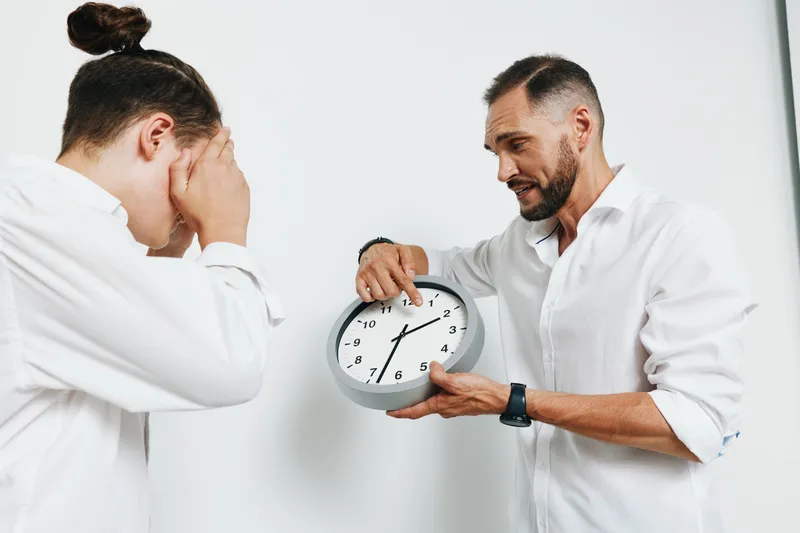 A professional man in a crisp shirt presents a clock while a colleague covers her face, showing time pressure and tense emotion against an isolated colored backdrop.