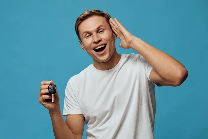 happy man holding car key smiling excited gesture listen attention standing on blue background casual white t-shirt studio portrait positive emotions concept