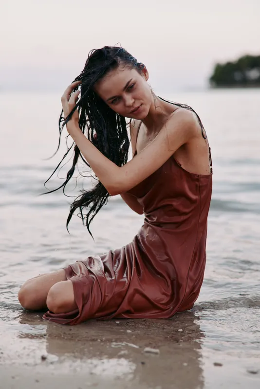 A solitary woman sits on the shallow shoreline, wet hair over a rust dress, gazing softly downward as gentle waves touch the sand, capturing calm solitude.