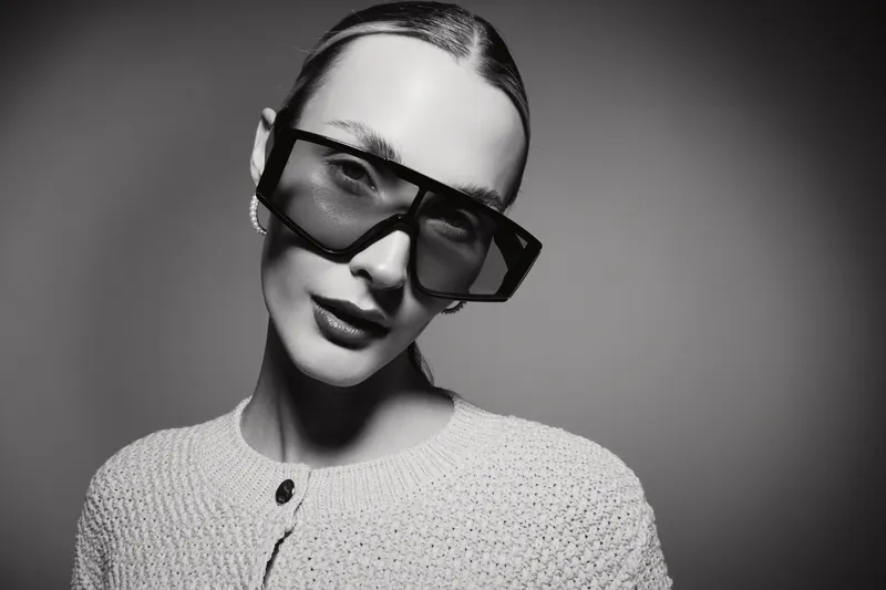 Portrait of a white young woman with light skin wearing oversized sunglasses and a knitted sweater. Studio black and white fashion concept with modern accessories and minimalistic style.