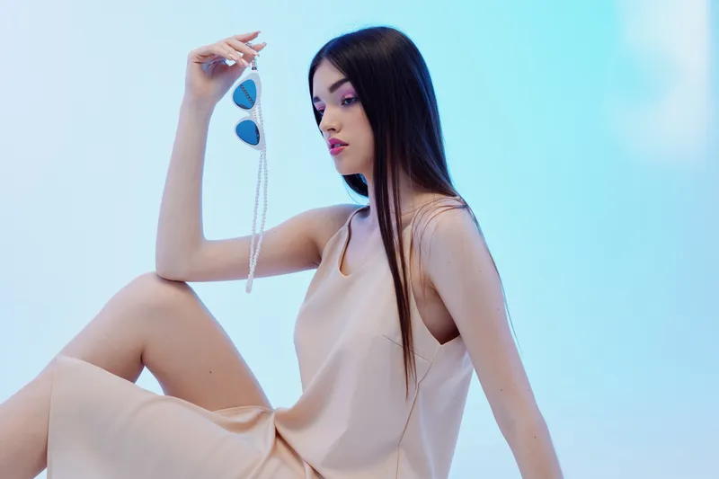 Young woman with long dark hair wearing a beige dress holds sunglasses in hand while sitting against a soft blue background. Concept of woman beauty fashion and style.