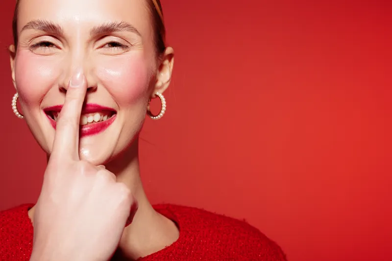 Smiling woman with red lipstick and hoop earrings touches her nose with finger. Close-up portrait on red background. Concept of happiness, beauty, and playful mood in studio lighting.