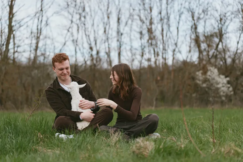 Cheerful couple outdoors with a playful goat kid in a meadow, candid moment of joy and interaction, warm natural light, relaxed posture, fresh air, vibrant colors, lifestyle scene