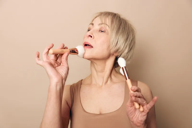 Mature woman with short blonde hair applies powder makeup using two brushes on beige background. Concept of beauty routine, skincare, self-care, and natural aging with elegance.