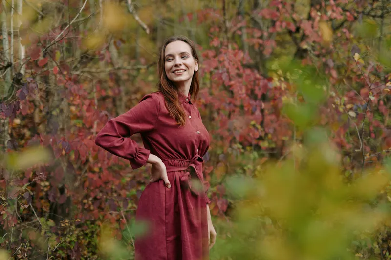A candid portrait of a smiling woman in a burgundy dress standing in a sunlit autumn grove, radiating warmth, ease, and authentic charm in natural light.