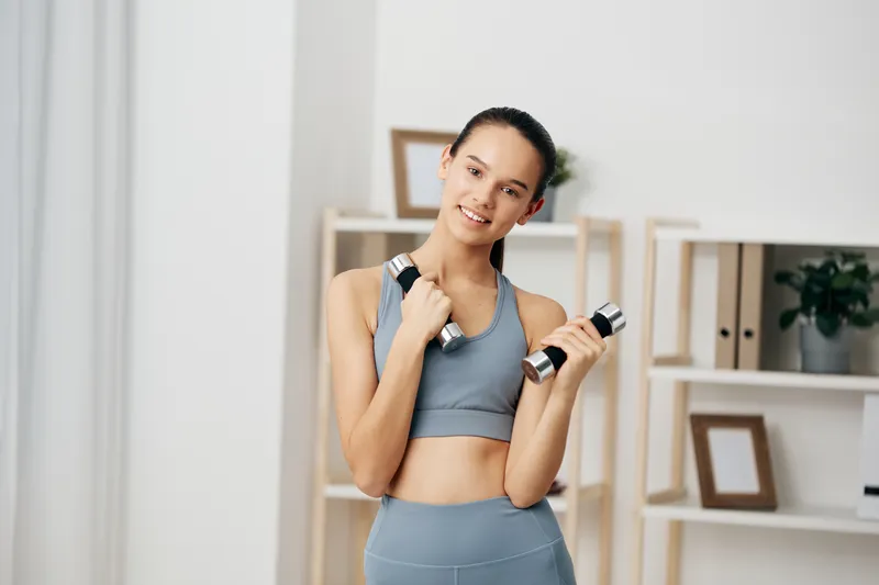 Fitness woman in athletic wear holding dumbbells for a home workout, showcasing strength, balance and confidence in a bright, minimal living space.