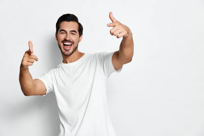 Happy cheerful young man wearing a white t-shirt, smiling widely and gesturing with hand, standing against a solid light background in a playful pose.