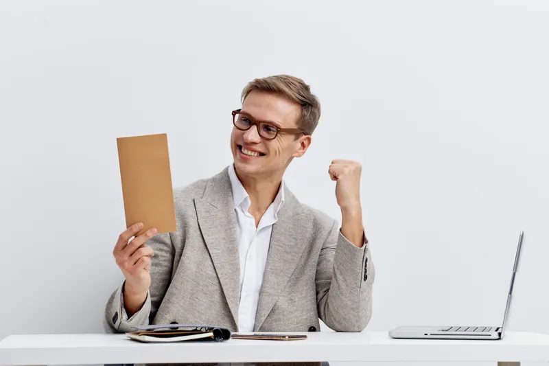 cheerful man celebrate success hold envelope wear glasses beige blazer white shirt sit at table with laptop concept digital nomad remote work freelance