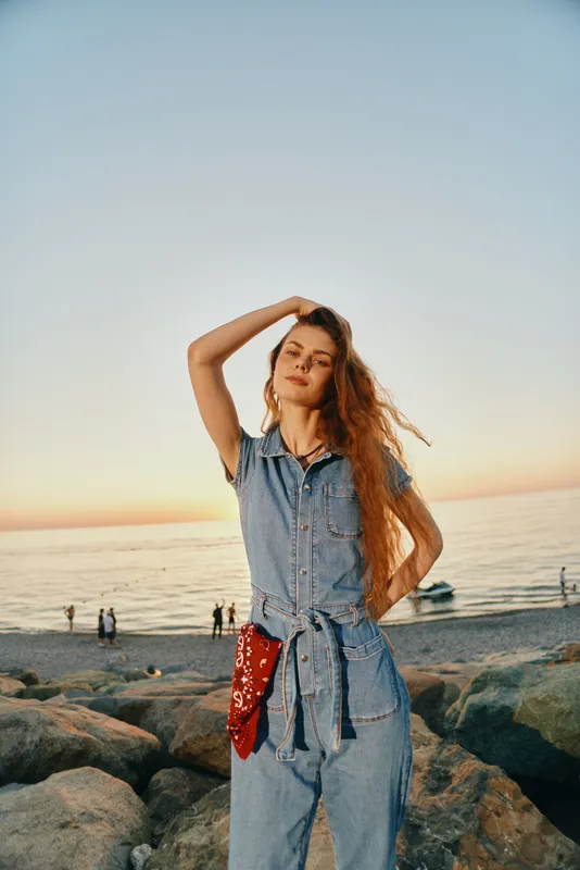 Lifestyle woman in modern boho-western denim street style stands confidently by the beach during sunset, showcasing film color warmth and casual summer vibe.