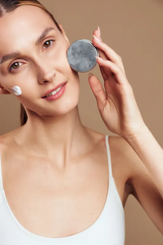 Young woman with fair skin applies cream on her cheek while holding a cosmetic jar near her face, smiling gently against a neutral background in a skincare routine scene.