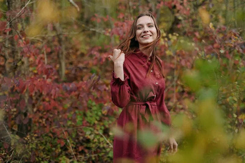 A young woman in a modest red dress smiles candidly among autumn foliage, a genuine portrait emphasizing authenticity, natural light, sincere expression and textured surroundings.