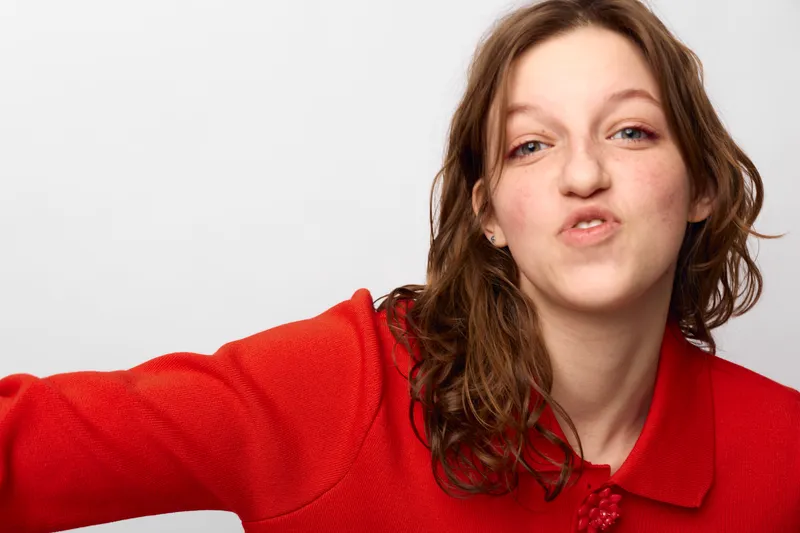 Young woman with curly brown hair wearing red sweater makes funny face with pursed lips on white background. Casual portrait with playful expression and natural lighting in studio setting.