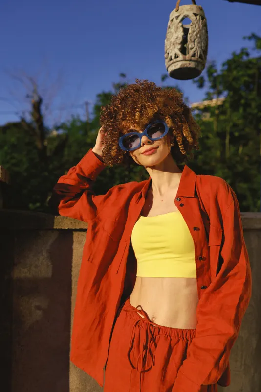Vibrant outdoor fashion portrait of a confident woman in a red jacket, yellow crop top, and orange pants, wearing sunglasses, posing in a sunlit urban setting with bold style