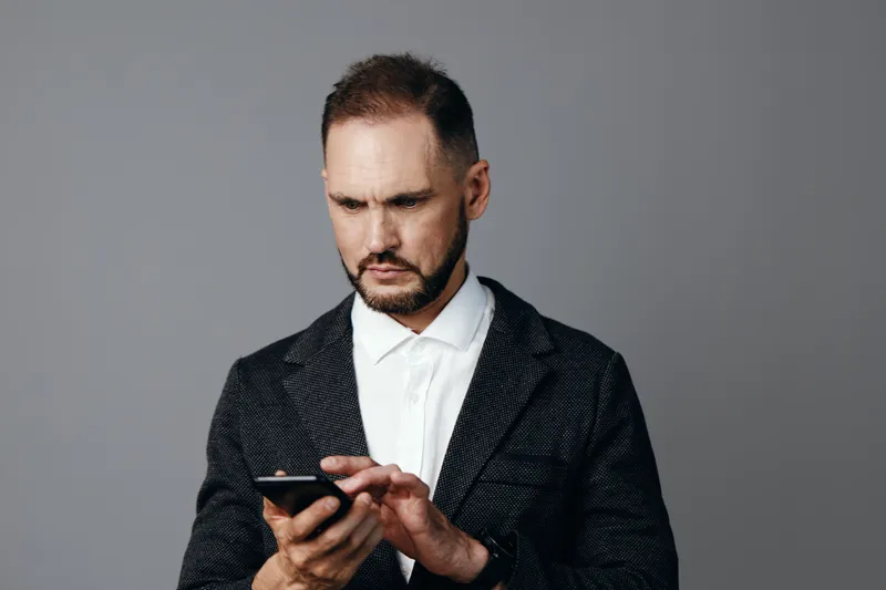 A business professional man in a dark blazer and white shirt stands before a neutral background, focused on his phone, projecting calm concentration and a ready, composed mood.