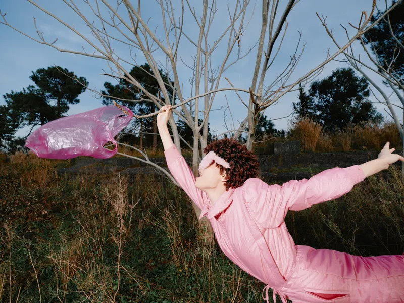 Pink clad performer leaps outdoors, reaching toward a bare tree with a flowing scarf, capturing a windy, playful moment of movement and whimsy in a natural field.