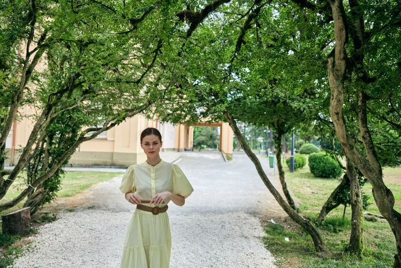Woman in a light yellow dress walking along a tree lined path in a tranquil park, exuding calm elegance and natural charm for a serene lifestyle scene