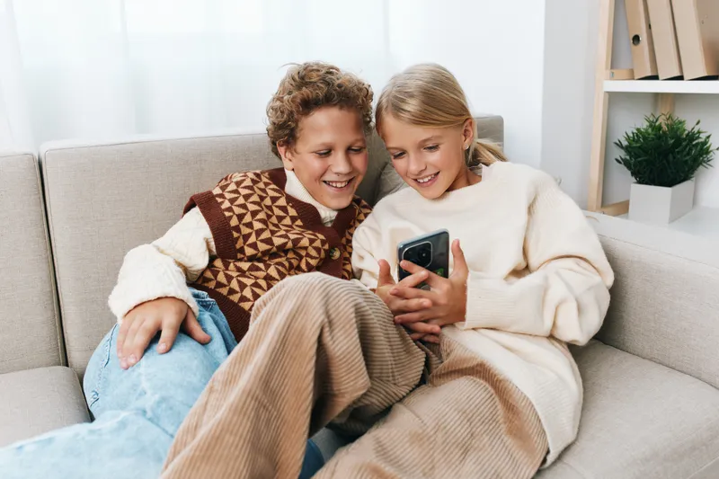 Two people sit close on a cozy sofa, smiling while sharing a smartphone, a warm moment of connection and comfort in a casual home setting.