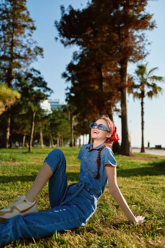 smiling woman relaxing outdoors in blue denim jumpsuit with sunglasses and red headscarf sitting on grass under sunny sky in park with tall trees