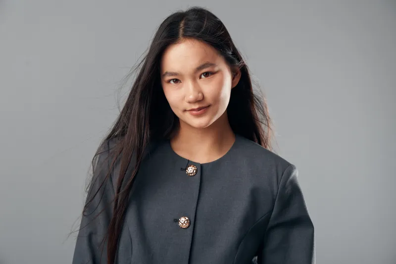 Young woman with long black hair wearing a gray jacket smiling gently in studio setting with plain background, casual portrait of confident Asian female person looking at camera.