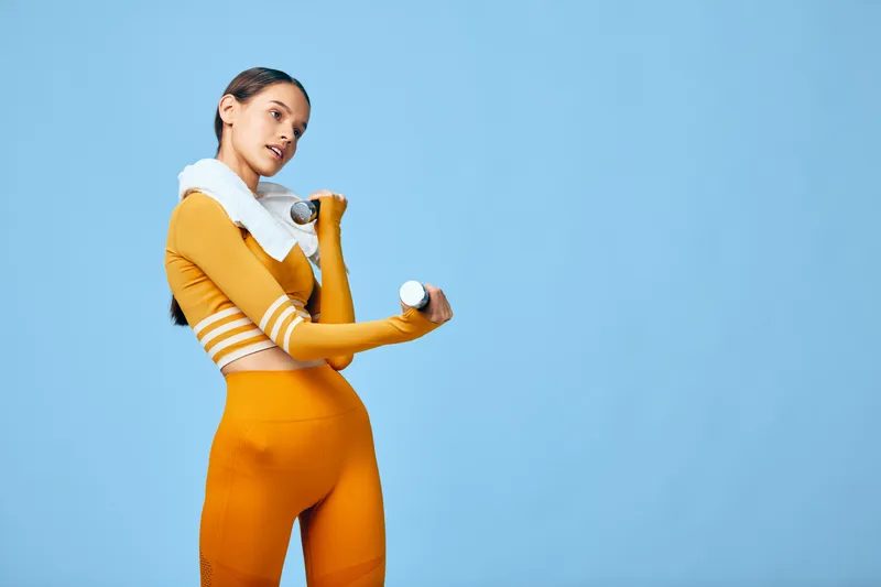 Fitness woman demonstrates workout and exercise with dumbbells in a bold orange outfit against a bright blue studio backdrop, showcasing strength, balance and confident motion.